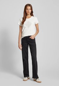 T-shirt bianca in cotone con stampa grafica geometrica, abbinata a jeans neri a gamba dritta e sneakers con accenti beige.