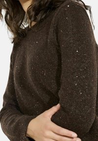 Femme aux cheveux ondulés foncés portant un pull en maille marron scintillant, tenant son bras gauche avec sa main droite sur un fond blanc.