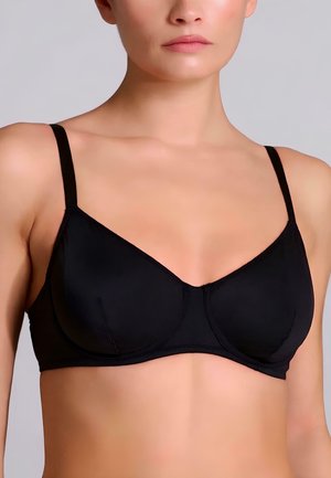 Donna che indossa un reggiseno nero leggermente imbottito con spalline sottili su uno sfondo chiaro e uniforme, che mostra la parte superiore del torso e il collo.