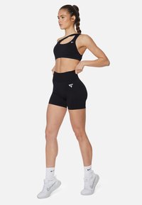 Sostén deportivo negro con diseño de tirantes asimétricos y logo; pantalones cortos de tiro alto negros a juego. Modelo con zapatillas Nike blancas.