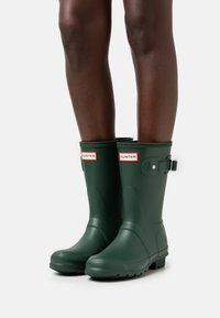 Botas de lluvia de goma verdes con una superficie texturizada, correa ajustable y detalles de logo en la parte superior. Diseño clásico de altura a la rodilla.