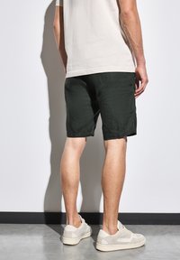 Tmavě zelené kraťasy z lehkého materiálu, krátké délky, s volným střihem. Oblečeno s béžovými teniskami a krémovým tričkem.