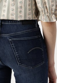 Mörka denimjeans med medelhög midja, försedda med två bakfickor och subtil vit sömnad i en böjd design. Svart läderlogotyp synlig.