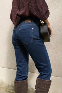 Persoon die blauwe jeans, bruine suède laarzen en een bruin top met patroon draagt en een kleine zwarte handtas tegen een beige muur vasthoudt.