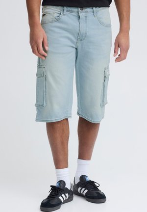 Mann trägt hellblaue Jeans-Cargo-Shorts, schwarzes T-Shirt, weiße Socken und schwarze Adidas-Sneaker und steht vor einem schlichten Hintergrund.