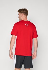 Rode T-shirt met korte mouwen van katoen, met een klein wit logo op de achterkant. Gepaarde met zwarte shorts met witte zijstrips.