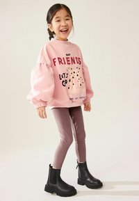 Felpa rosa chiaro con maniche a sbuffo, caratterizzata da stampe testuali e grafiche; abbinata a leggings marroni e stivaletti neri alla caviglia.