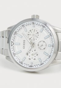 Reloj de pulsera Guess plateado con esfera blanca, tres subesferas que indican el día, la fecha y la hora en formato de 24 horas, y marcadores y manecillas luminosos.