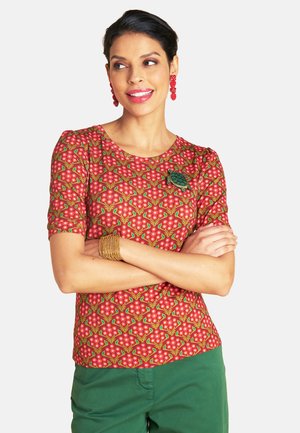 Frau mit dunklem Haar, die ein rotes, geblümtes Oberteil und grüne Hosen trägt, rote Ohrringe, ein goldenes Armband und eine grüne Schildkrötenbrosche, mit verschränkten Armen, seitlich lächelnd.