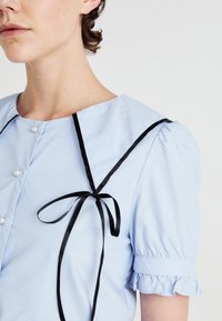 Lys blå bluse med perleknapper, korte puffærmer med flæser, og sort bånddetalje der danner en sløjfe nær kraven.