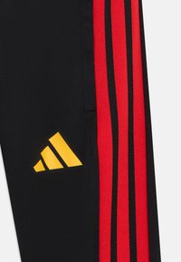 Calças desportivas pretas com riscas laterais vermelhas e detalhes pretos. Logótipo amarelo exibido próximo à área do bolso. Textura lisa.