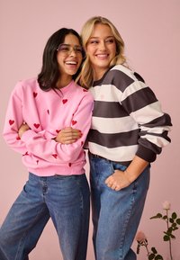 Dve usmievajúce sa ženy stoja blízko seba; jedna má na sebe ružovú mikinu so vzorom srdiečok a okuliare, druhá čierno-bielu pruhovanú mikinu.
