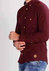 Man justerar ärmen på en långärmad vinröd skjorta med beige knappar, iklädd blå jeans.