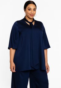 Marineblauwe top van gladde stof met een keyhole-ontwerp bij de halslijn en gedrapeerde mouwen. Gecombineerd met bijpassende broek voor een gecoördineerde look.