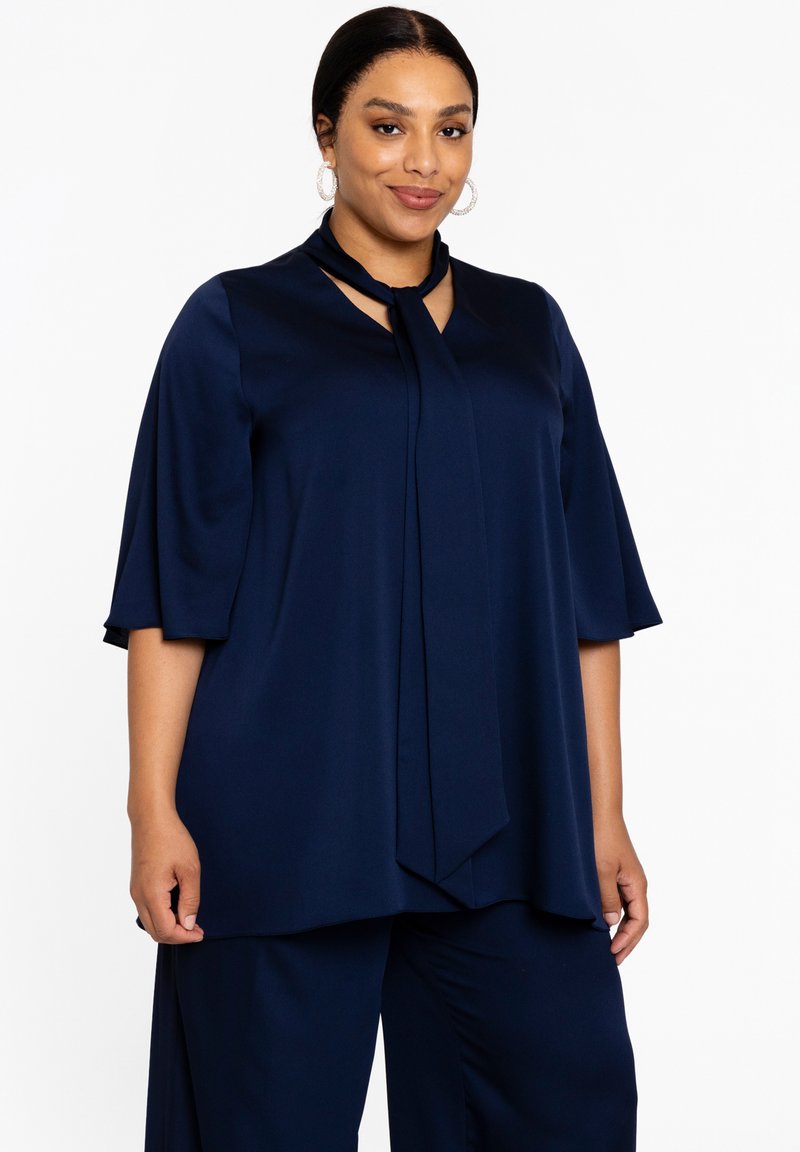 Marineblauwe top van gladde stof met een keyhole-ontwerp bij de halslijn en gedrapeerde mouwen. Gecombineerd met bijpassende broek voor een gecoördineerde look.