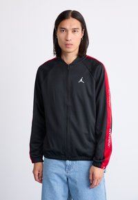 Black zip-up jacket with red side stripes, featuring a white logo on the chest and “JORDAN” text along one sleeve. Smooth fabric texture.