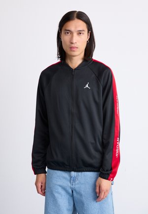 Sort zip-jakke med røde side-striber, der har et hvidt logo på brystet og "JORDAN" tekst langs en ærme. Glat stofstruktur.
