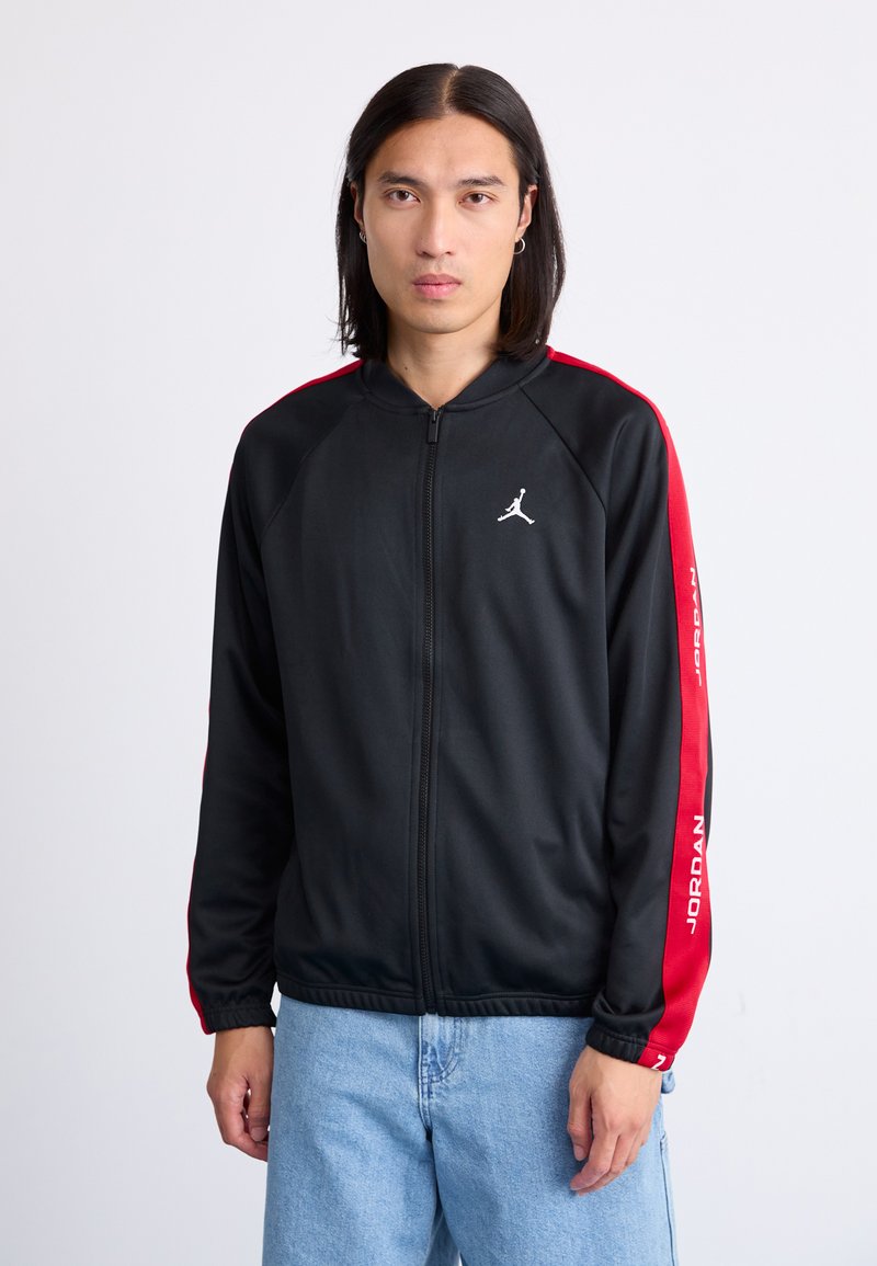 Black zip-up jacket with red side stripes, featuring a white logo on the chest and “JORDAN” text along one sleeve. Smooth fabric texture.
