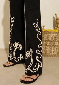 Black wide-leg pants with white embroidered palm tree and curvy designs. Textured fabric and worn with black sandals featuring gold accents.