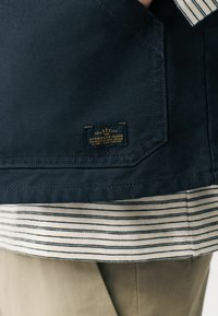 Marineblauwe jas met gestreepte onderzoom, voorzien van een gestikte patch met het label "STANDARD ISSUE" in goud en een textuurafwerking.