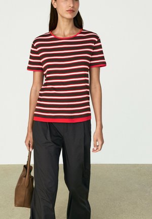 Frau trägt ein rot, pink und schwarz horizontal gestreiftes kurzärmeliges Shirt mit schwarzen Hosen und hält eine braune Handtasche an ihrer Seite.