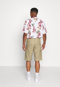 Kurzes weißes Hemd mit rot-gelbem Blumenmuster, kombiniert mit knielangen khakifarbenen Cargo-Shorts und weißen Sneakers.