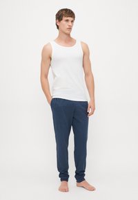Jonge man die blootsvoets staat, gekleed in een witte mouwloze shirt en blauwe casual broek, tegen een plain witte achtergrond.