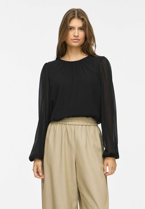 Jeune femme aux longs cheveux bruns portant un chemisier noir transparent à manches longues et un pantalon beige taille haute à jambes larges, debout devant un fond blanc.