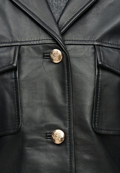 Μαύρο δερμάτινο μπουφάν με δύο χρυσά ανάγλυφα κουμπιά και τσέπες με καπάκι πάνω από γκρίζο πλεκτό πουλόβερ.