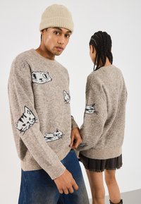 Beige gestrickter Pullover mit verschiedenen Katzenillustrationen in Schwarz und Weiß. Lockere Passform mit gerippten Bündchen und Saum. Trägt er mit blauen Jeans und einem Rock.