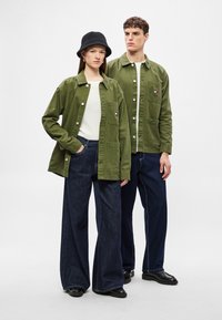 Δύο μοντέλα φορούν μπουφάν ελιάς από ανθεκτικό ύφασμα, σε συνδυασμό με φαρδιά παντελόνια από σκούρο τζιν. Τα μπουφάν έχουν κουμπιά και τσέπες.