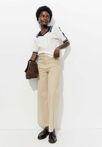 Polo shirt en tricot blanc avec des accents bleu marine, associé à un pantalon large beige. Tient un sac à main à motif léopard et porte des chaussures brillantes sombres.