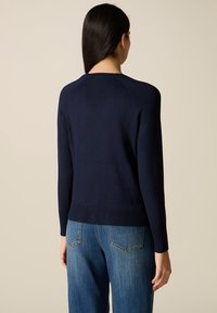 Femme aux longs cheveux bruns portant un pull bleu marine et un jean bleu, vue de dos sur un fond beige uni.