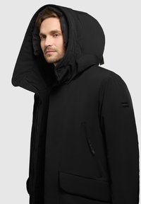 Sort parka-jakke med en stor, polstret hætte, lynlåslomme i siden og en struktureret overflade. Har minimal branding på ærmet.