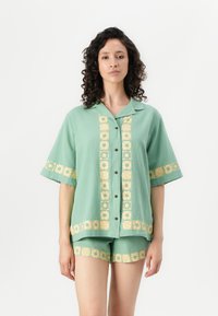 Groene shirt met korte mouwen en gele bloempatronen, met knoopsluiting aan de voorkant. Past bij groene shorts met vergelijkbare ontwerpen. Lichtgewicht stof.