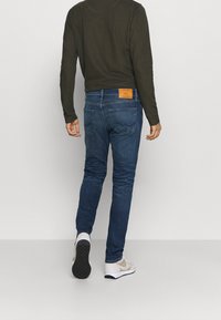 Blå denimjeans med raka ben och en brun läderetikett bak, kombinerade med en mörk långärmad tröja och ljusgrå sneakers.