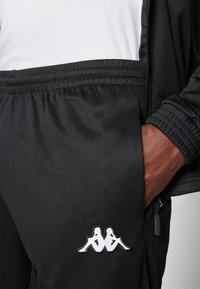 Set de trening sportiv negru, compus dintr-o jachetă cu fermoar și pantaloni cu talie elastică, având logo-ul alb Kappa. Textură netedă, din material sintetic.