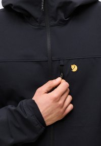 Hånd som drar glidelåsen på en svart jakkelomme med liten gul reve-logo på brystet, viser frontglidelås og hettekrage.