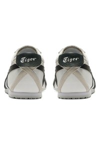 Hvide athletic sko med struktureret læder og ruskindsdetaljer, udstyret med sorte og beige krydsguider og en sort heel tab med brand.