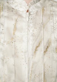 Gros plan d’un vêtement blanc en soie avec des motifs marbrés dorés et une fermeture éclair blanche centrée, avec un col court à volants.