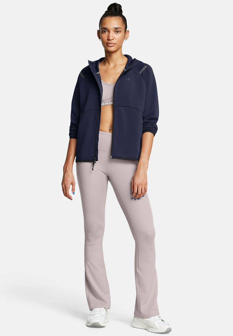 Veste à capuche zippée navy sur un soutien-gorge de sport gris clair et des leggings roses évasés, associés à des baskets blanches, avec une texture lisse.