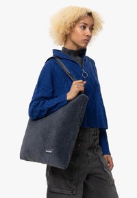 Borsa tote in fleece grigio scuro tenuta da una persona, caratterizzata da una texture morbida e un'etichetta con logo. La borsa ha una forma rettangolare semplice con una tracolla nera.