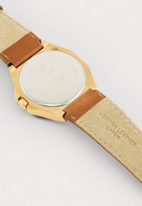 Orologio con cassa rotonda in acciaio inossidabile color oro. Cinturino in vera pelle marrone, texture liscia, dettagli in rilievo e finitura lucida.