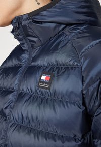 Námornícka puffer bunda vyrobená z lesklého prešívaného materiálu s výraznými sekciami, so zipsom, vysokým golierom a označením s logom na hrudi.