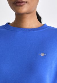 Blå bomull crewneck genser med ribbestrikket krage og brodert logo på brystet, med en jevn tekstur og avslappet passform.