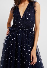 Laivastonsininen tüllimekko, jossa on syvä V-mallinen kaula-aukko, puffat hihat, vartalonmyötäinen yläosa ja kerroksellinen hame, jossa on monivärisiä pilkkuja.