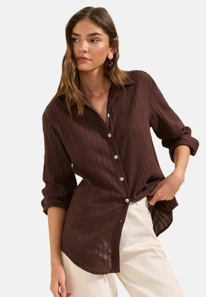 Femme portant une chemise marron texturée à boutons avec manches retroussées et un pantalon beige clair, posant avec une main dans la poche devant un fond uni.