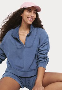 Modrý sweatshirt z bavlněné směsi s polo zipem, žebrovanými manžetami a odpovídajícími šortkami. Nosí se s světle růžovou baseballovou čepicí.