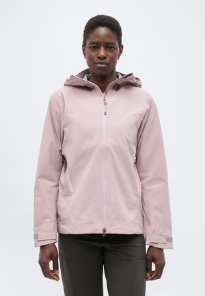 Persoană văzută din față, purtând o jachetă cu glugă roz deschis cu fermoar și pantaloni verde închis, pe un fundal alb simplu.