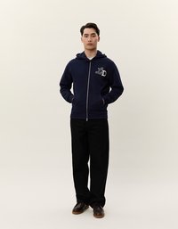 Felpa con zip di colore blu navy e coulisse, logo "L&D" bianco sul petto, abbinata a pantaloni neri e scarpe marroni. Tessuto dalla texture liscia.